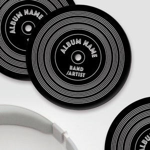 Puede incluir: Tres posavasos de vinilo negro con texto blanco que dice "Album Name", "Band/Artist". Un par de auriculares blancos est&aacute; en primer plano.