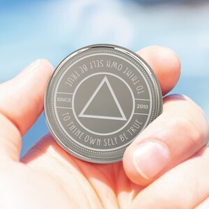 May include: A silver coin with a triangle in the center and the text "To thine own self be true" around the edge. The coin also has the text "Since 2010" on the bottom right and "To thine own self be true" on the top left.