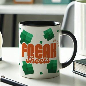 May include: White ceramic mug with a black rim and handle. The mug has a green and orange design with the text "Freak Sheets" and "In The" printed on it.