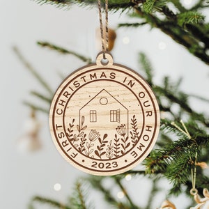 May include: A round wooden ornament with the text "First Christmas in Our Home 2023" and a house with a window and a door, surrounded by flowers.