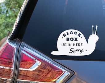 May include: A white snail sticker on a car's back window. The snail has a black circle on its shell with the text "BLACK BOX UP IN HERE Sorry..."