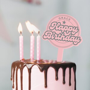 May include: A pink cake with chocolate frosting and three lit candles. A pink cake topper with the text "GRACE Happy Birthday" is on top of the cake.