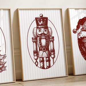 May include: Three framed Christmas art prints. The prints feature a Christmas tree, a nutcracker, and Santa Claus, all in a vintage style. The artwork is in a deep red color on a white background with vertical stripes. The frames are a light wood color.