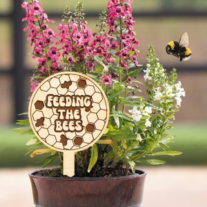 K&ouml;nnte beinhalten: Ein Holzschild mit dem Text "Feeding the Bees" umgeben von einem Wabenmuster. Das Schild steckt in einem Topf mit rosa und wei&szlig;en Blumen, in der N&auml;he fliegen zwei Hummeln.