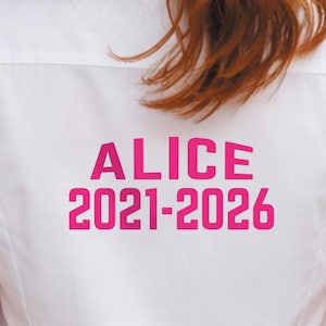 Pu&ograve; includere: Camicia bianca abbottonata con il nome "ALICE" e gli anni "2021-2026" stampati in rosa acceso sul retro. La camicia &egrave; realizzata in tessuto liscio. L'immagine mostra il retro della camicia.