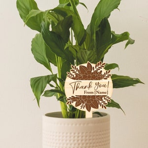 May include: A wooden plant stake with a brown floral design and the text "Thank you! From [Name]" in a white font.