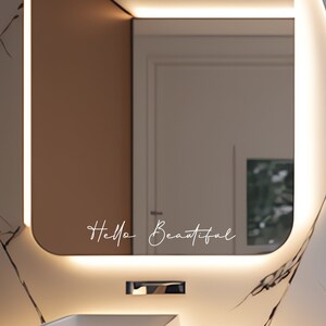 Puede incluir: Un espejo de ba&ntilde;o moderno con un borde de luz LED blanco y el texto "Hello Beautiful" escrito en la superficie del espejo.