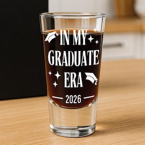 May include: A clear shot glass filled with a dark liquid, featuring the text "IN MY GRADUATE ERA - 2026" in white. The glass is set on a wooden surface, with a black box in the background.
