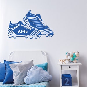 May include: Blue and white football boot wall decal with the name Alfie on a white wall in a bedroom setting. The decal is above a white bed with blue bedding and pillows. A white nightstand with a drawer and a shelf is next to the bed. The shelf has a blue box with the number 2 on it. A white basket with white and blue balls is on the nightstand.