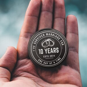 Puede incluir: Un recuerdo redondo en blanco y negro con el texto "I'VE SURVIVED MARRIAGE FOR... 10 YEARS ESTD 2014 ONE DAY AT A TIME" y dos anillos entrelazados. El recuerdo se sostiene en una mano abierta.