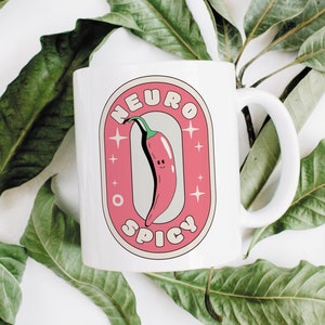 Puede incluir: Taza de cer&aacute;mica blanca con un gr&aacute;fico rosa y blanco de un chile rojo con una cara sonriente. El gr&aacute;fico dice "Neuro Spicy" en una fuente retro.