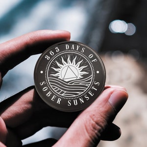 May include: A black metal coin with a white design. The coin features a sun with a triangle inside, waves, and the text "365 Days of Sober Sunsets".