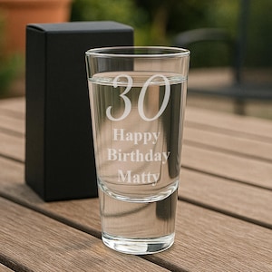 May include: A clear shot glass with the number "30" and the words "Happy Birthday Matty" etched into the glass. The glass is filled with a clear liquid and sits on a wooden surface next to a black gift box.