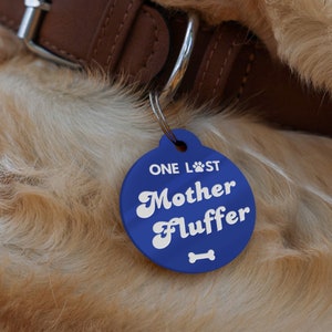 Op de afbeelding: Blauwe hondenhanger met witte tekst die "ONE LOST Mother Fluffer" luidt, met een bot-icoon onder de tekst.
