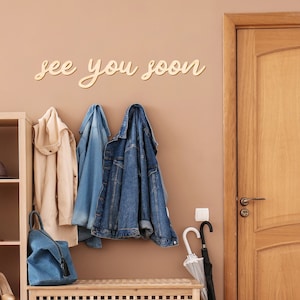 Peut inclure: Une entr&eacute;e de maison avec un banc en bois, un porte-manteau et une porte en bois. Le mur est d&eacute;cor&eacute; de la phrase "see you soon" en &eacute;criture cursive. Des manteaux et un sac en jean sont accroch&eacute;s au porte-manteau, et un parapluie est appuy&eacute; contre le mur.
