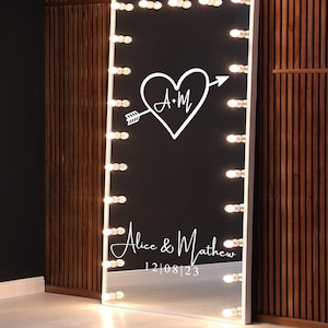 May include: A full-length mirror with a white frame and a row of light bulbs around the edge. The mirror has a black background with a white heart and arrow design with the initials "A+M" inside the heart. The text "Alice & Matthew" and the date "12|08|23" are written below the heart.