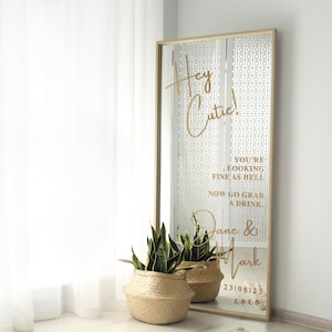 May include: A gold mirror with a geometric pattern and the text "Hey Cutie! You're looking fine as hell. Now go grab a drink. Jane & Mark 23108123 xoxo" on it. A potted plant is in front of the mirror.