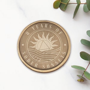 May include: A gold-toned round coin with the words "3 Years of Sober Sunsets" engraved around the edge. The center of the coin features a stylized sun with a triangle inside, and waves below.