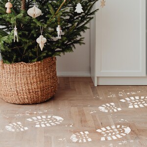 May include: A Christmas tree decorated with white ornaments sits in a wicker basket on a wood floor. White footprints are painted on the floor.