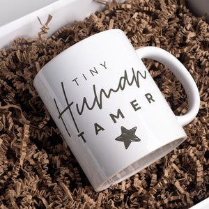 May include: White ceramic mug with black text that reads "Tiny Human Tamer" and a black star. The mug is sitting in brown crinkled paper.