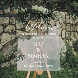 May include: A clear acrylic welcome sign with white text. The sign reads "Welcome to our Roka Ceremony Raj & Simran 08 November 2023".