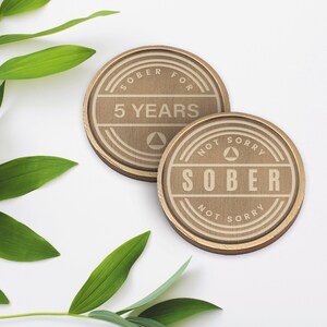 May include: Two round wooden coasters with gold trim. One coaster says "Sober for 5 Years" and the other says "Not Sorry Sober Not Sorry".