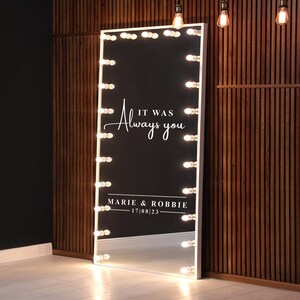 May include: A white vanity mirror with a black background and white text that reads "It was always you" and "Marie & Robbie - 17/08/23". The mirror has a white frame and is surrounded by a row of light bulbs.