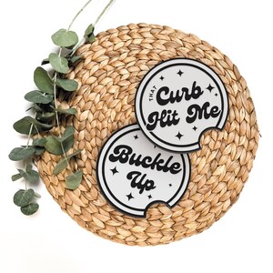 May include: Two round coasters with black text on a white background. The coasters are on a woven straw mat. The text on the coasters reads "That Curb Hit Me" and "Buckle Up".