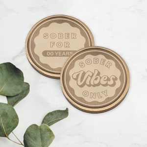 K&ouml;nnte beinhalten: Zwei runde Metallmarken in Goldfarbe mit erh&ouml;htem Rand. Die erste Marke lautet "SOBER FOR 00 YEARS". Die zweite Marke lautet "SOBER Vibes ONLY".