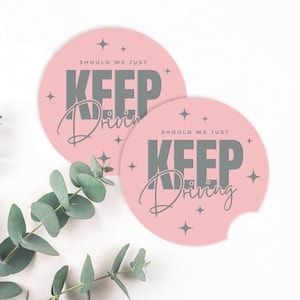 May include: Two pink round coasters with a grey text overlay that reads "Should we just keep driving". The coasters have a cut-out shape for a beverage.