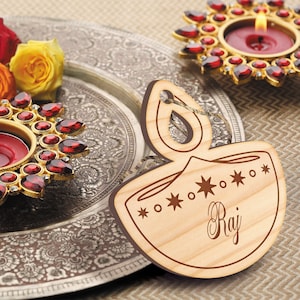 K&ouml;nnte beinhalten: Ein h&ouml;lzerner Anh&auml;nger in Diya-Form mit dem eingravierten Namen "Raj". Der Anh&auml;nger ist mit Sternen und Kreisen am Rand verziert. Er liegt auf einem silbernen Tablett mit rotem und goldenem Design.