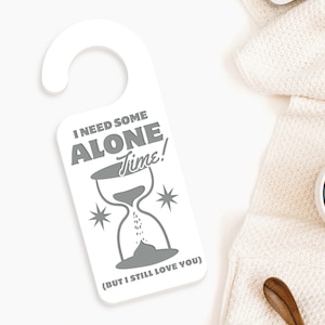 May include: White door hanger with a grey hourglass and the text "I need some alone time! (But I still love you)".