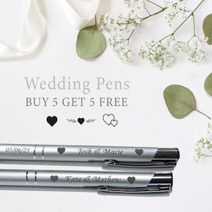 May include: Two silver pens with black accents and personalized engravings. The top pen reads "05/06/25 Josh & Macie" and the bottom pen reads "Kate & Matthew". The text "Wedding Pens BUY 5 GET 5 FREE" is displayed above the pens.  Four heart icons are displayed below the text.