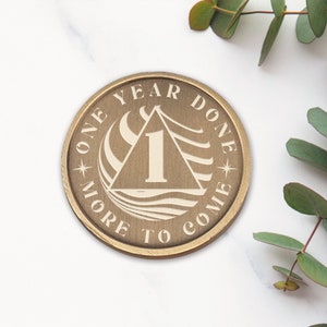 Op de afbeelding: Een gouden munt met de woorden "One Year Done - More To Come" en het getal "1" in een driehoek. De munt ligt op een wit marmeren oppervlak.