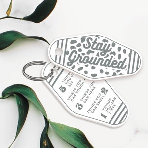 May include: White keychain with a grey motel key design. The keychain has the text "Stay Grounded" and a list of five things you can see, touch, hear, smell, and taste.
