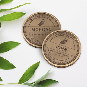 Puede incluir: Dos monedas de decisi&oacute;n de madera con texto grabado. Una moneda dice "MORGAN" y la otra "JOHN". Ambas monedas tienen el texto "FLIP TO DECIDE" y "DECISION CO" alrededor de los bordes. Hojas verdes en el fondo.