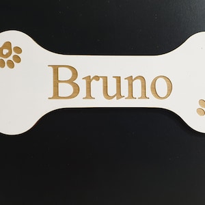 May include: White wooden dog bone with the name "Bruno" engraved in gold lettering. The bone has two gold paw prints on either side of the name.