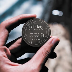 Puede incluir: Un token redondo negro con letras doradas que dice "YOUR SOBRIETY IS A BIG DEAL I SEE YOU & I AM SO PROUD OF YOU".