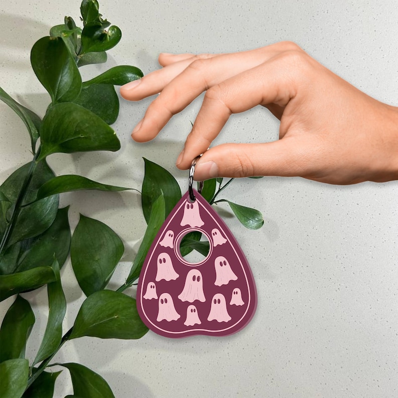 May include: A burgundy-colored, heart-shaped, wooden charm with a hole in the center and a pink ghost pattern. The charm is attached to a silver key ring.