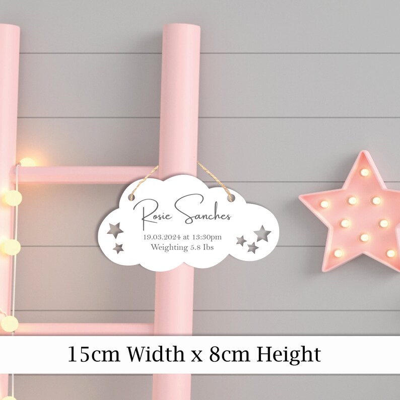 May include: White wooden cloud-shaped sign with the text "Rosie Sanchez" and the date "19.03.2024 at 13:30pm" and "Weighting 5.8 lbs". The sign is hanging from a pink ladder with a pink star light behind it. The sign measures 15 cm wide by 8 cm high.