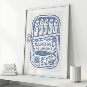 May include: A framed print featuring a blue illustration of a sardine can with the text "SARDINE AL LIMONE." The can contains six stylized fish. Decorative elements include lemons and leaves. The print is displayed on a white shelf with a stack of books and a white vase.