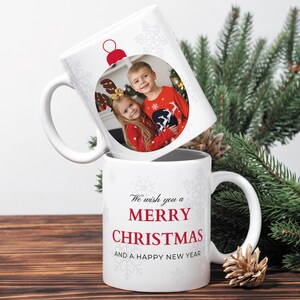 May include: Two white ceramic mugs, one stacked on top of the other. The top mug features a photo of two children in a Christmas ornament design. The bottom mug has the text "We wish you a Merry Christmas and a Happy New Year" in red.