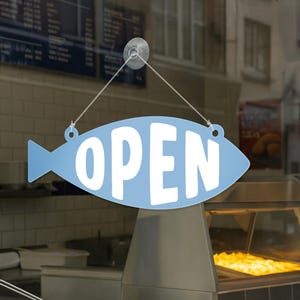 May include: A light blue, fish-shaped sign with the word "OPEN" in white lettering. The sign is hanging in a window, indicating a business is open. The sign is suspended by a clear suction cup and string.