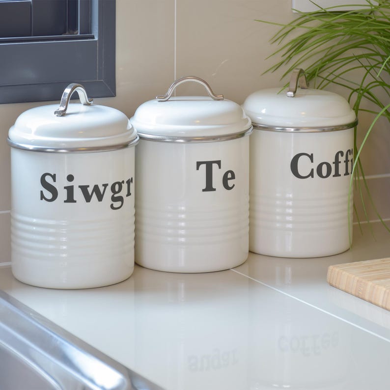 May include: Three white kitchen canisters with silver trim and handles. The canisters are labeled "Siwgr", "Te", and "Coff". The canisters have a ribbed design and are sitting on a white countertop.