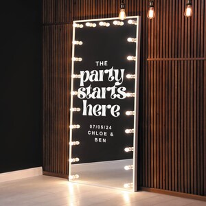May include: A white, full-length mirror with a white frame and a black background. The mirror has a row of light bulbs around the perimeter. The mirror has the text "THE party starts here" and "07/05/24 CHLOE & BEN" written on it.