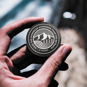 May include: A black round coin with white text and a mountain range design. The text reads "Make the comeback stronger than the setback - 6 years".