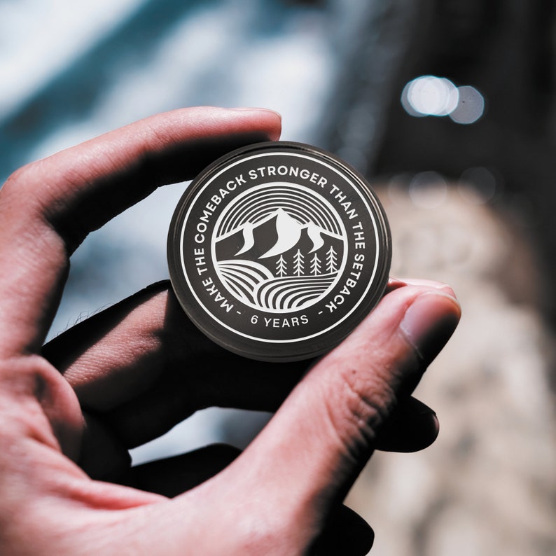 May include: A black round coin with white text and a mountain range design. The text reads "Make the comeback stronger than the setback - 6 years".