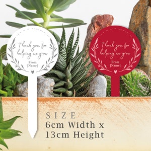 May include: Two garden stakes with a round white and red circle. The white stake has gray text that says "Thank you for helping me grow. From [Name]" and the red stake has pink text that says "Thank you for helping me grow. From [Name]" Both stakes are 6 cm wide and 13 cm high.
