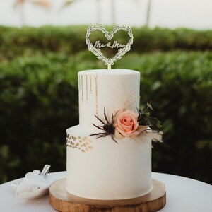 May include: A two-tiered white wedding cake with gold accents and a floral heart topper that says "Mr. & Mrs.". The cake is decorated with a single pink rose and greenery.