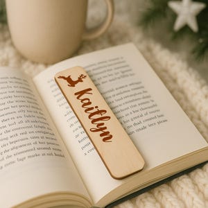 May include: A wooden bookmark with a reindeer graphic and the name "Kaitlyn" in a brown script font. The bookmark is resting on the pages of an open book. The bookmark is approximately 6 inches long.